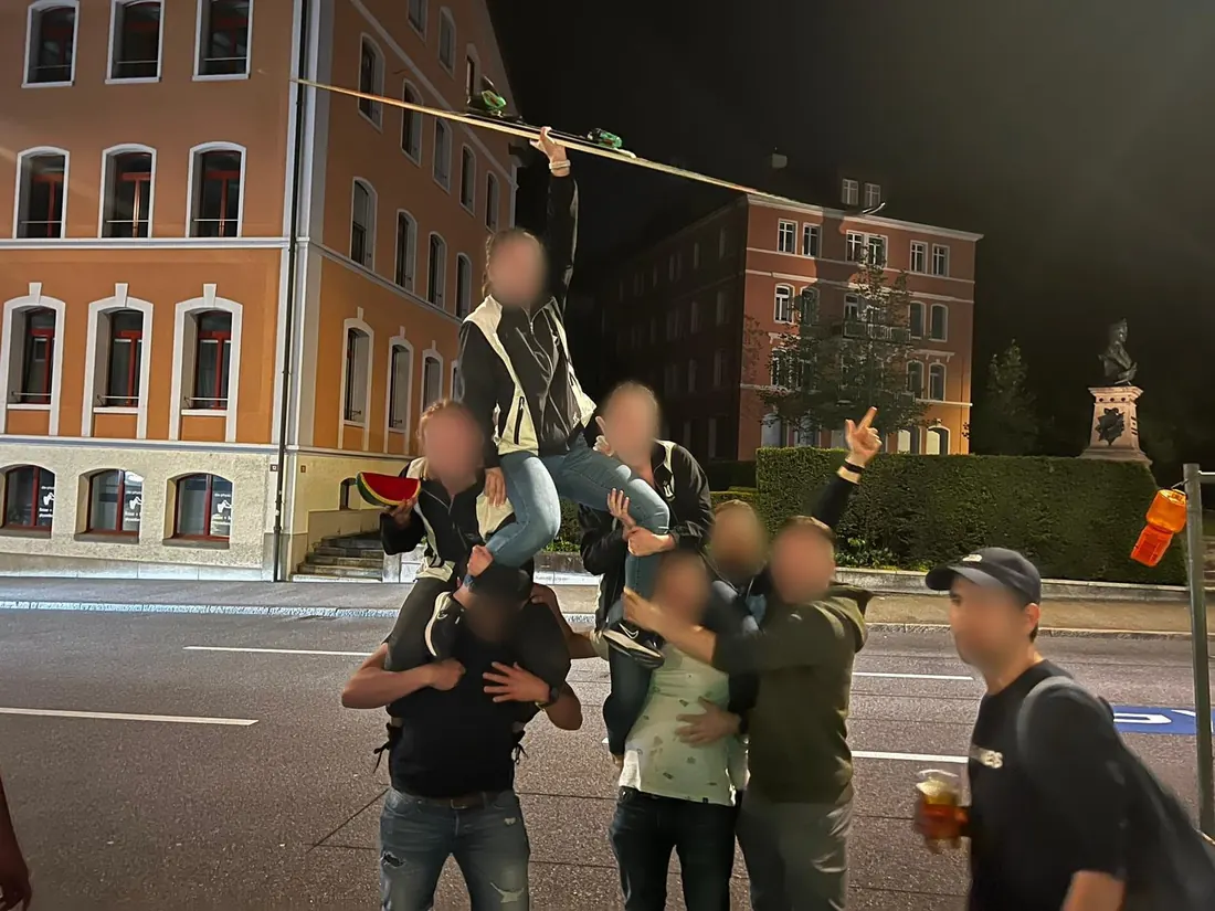 Freunde bilden nachts auf der Strasse eine Menschenpyramide, oben hält jemand einen Ski hoch.